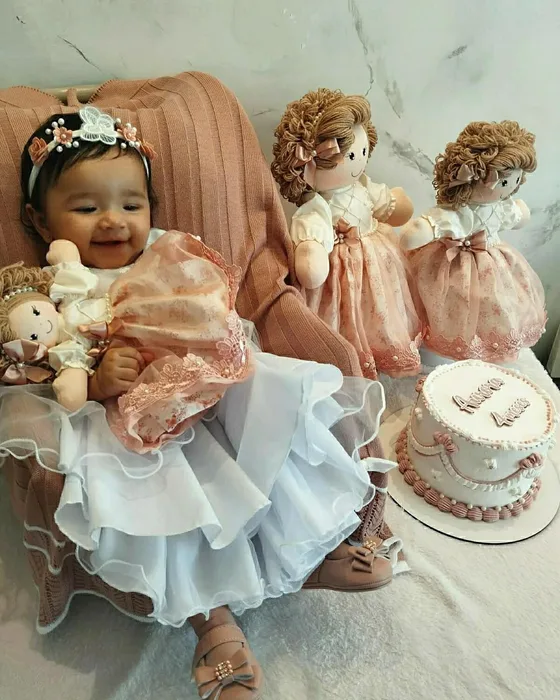 Bebê de vestido azul no aniversário, cercada de bonecas de pano de vestido rosé
