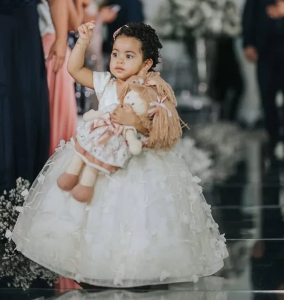 Menina de vestido branco na cerimônia de casamento, abraçada a uma boneca de pano