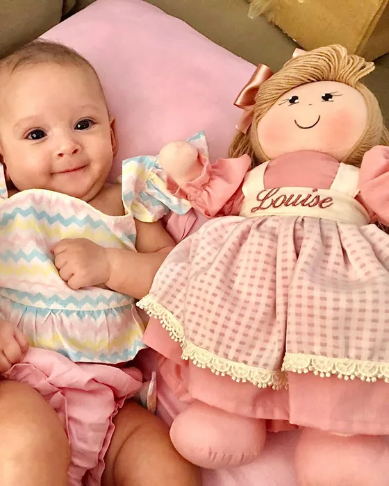 Bebê sorrindo ao lado de uma boneca de pano com o nome Louise bordado