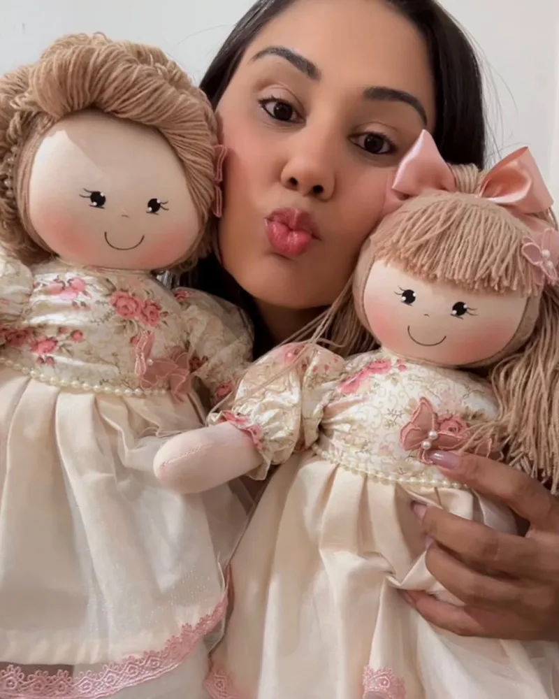 Ranielle mandando um beijo entre duas bonecas de pano de vestido florido