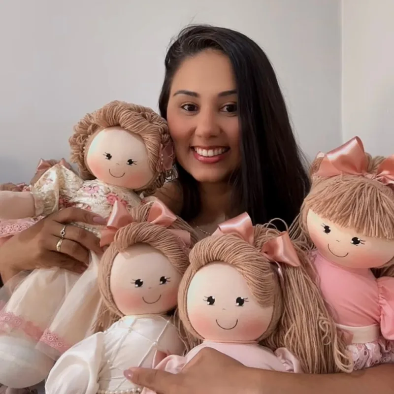 Ranielle sorrindo, abraçada a cinco bonecas de pano de laço rosé