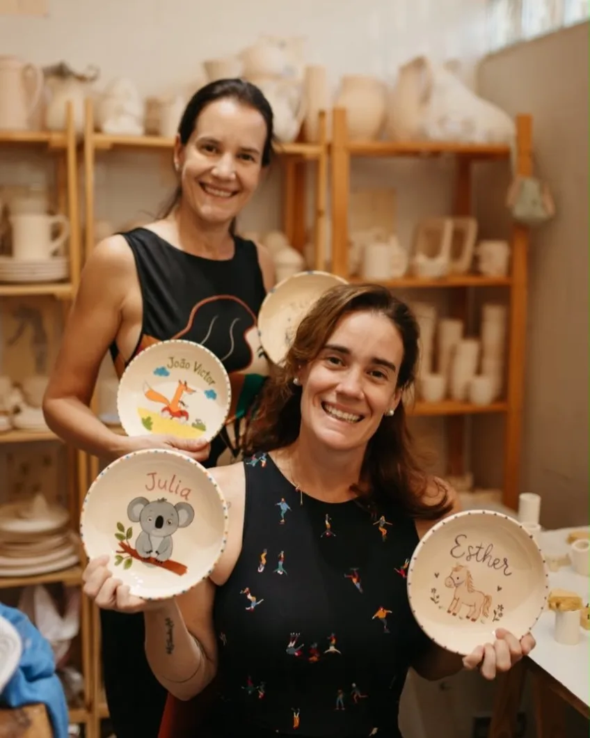 As irmãs Talita e Karina sorrindo no ateliê, mostrando pratinhos infantis pintados à mão, com estantes de peças ao fundo