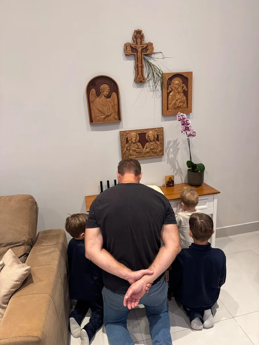 Pai ajoelhado com os três filhos diante do oratório da casa